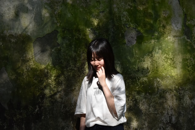 A woman in a white shirt smiles shyly and stands in front of a moss-covered wall. 
