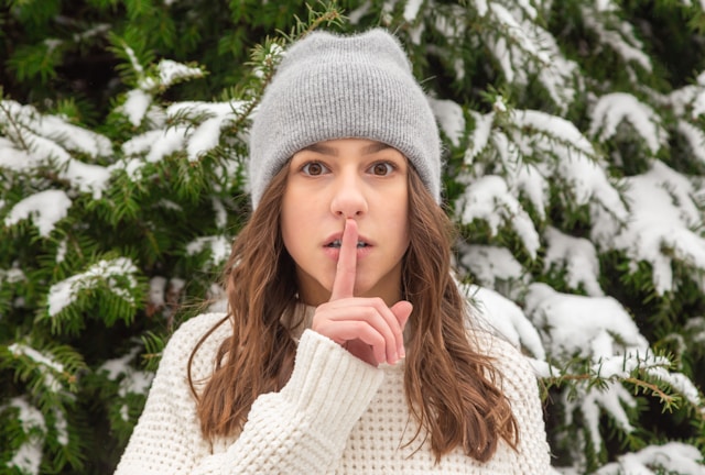 A young woman in a knit cap stands in front of a snow-covered tree and holds her index finger in front of her lips.