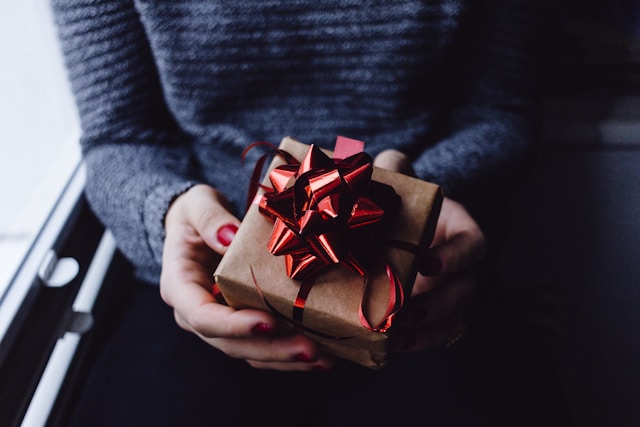 A person holds a gift box wrapped in brown paper and a red bow. 

