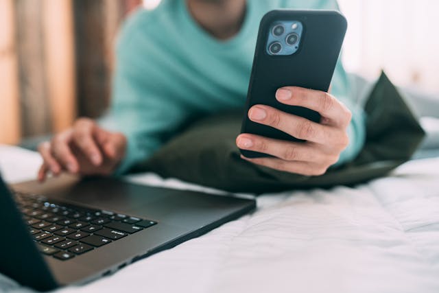 A man in a sweater types on a phone while using a laptop.
