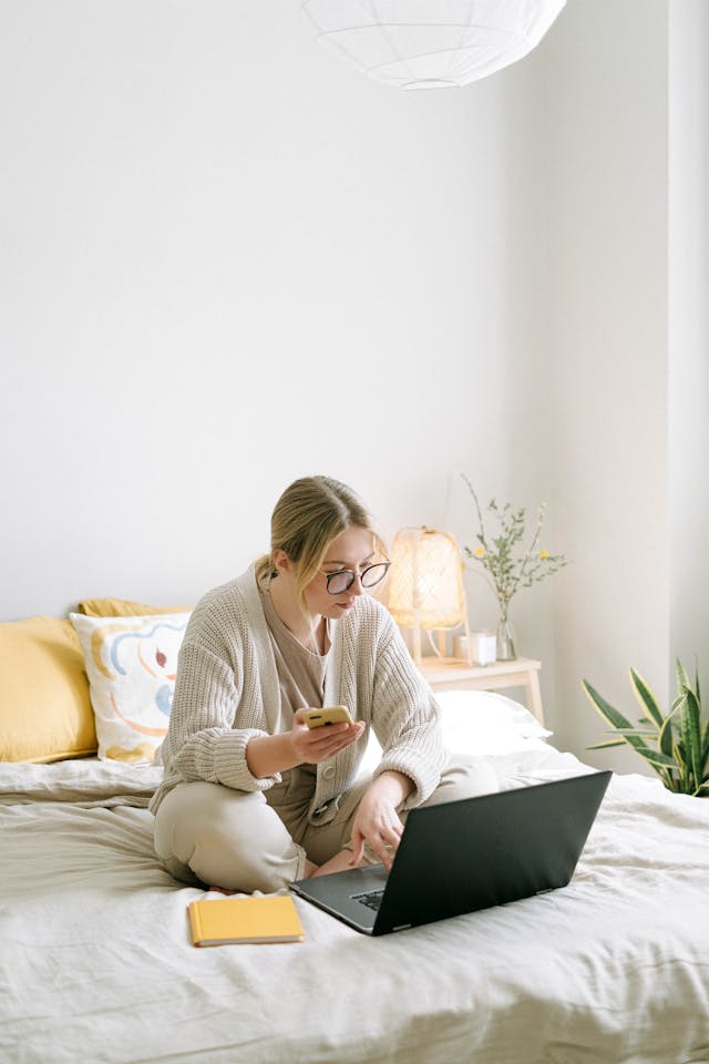 A woman sits on a bed using her phone and laptop.
