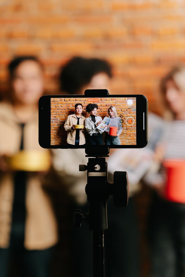 A camera phone records a video of three friends standing against a brick wall.
