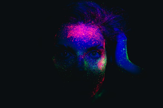 A face covered in different colors of glow-in-the-dark paint hides in the shadow. 
