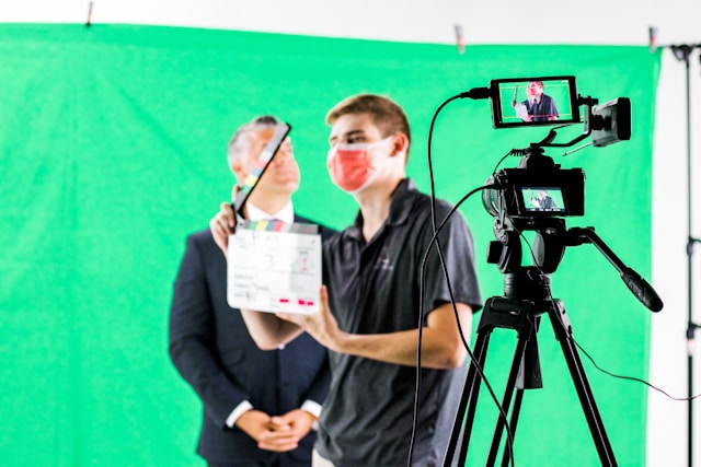 A man holds a clapperboard next to a video camera, and another man in a suit stands in front of a green background. 
