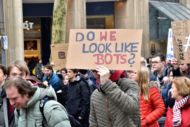 A person in a crowd holds up a cardboard sign that says, “Do we like bots?”