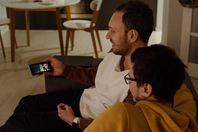 A couple sits on a couch and watches videos on a phone. 
