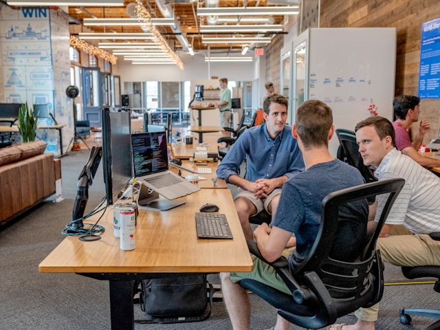 A group of professionals huddles in front of their computers and discusses their advertising strategy. 
