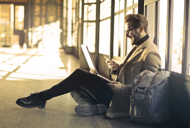 A man in a suit jacket sits on the floor and uses his phone and laptop at the same time. 
