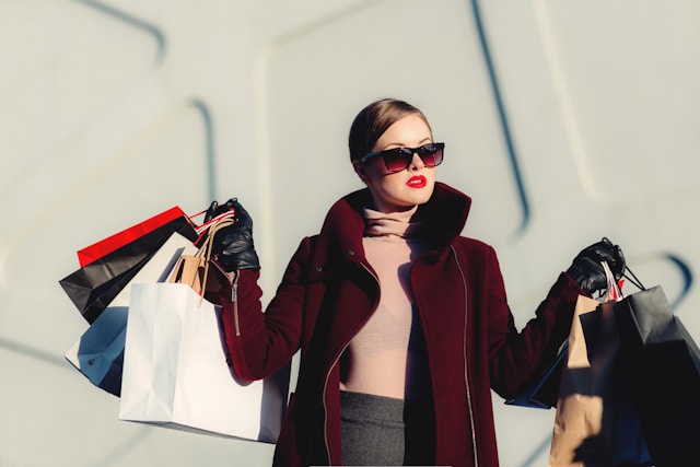 A woman wearing sunglasses and a coat carries lots of shopping bags. 
