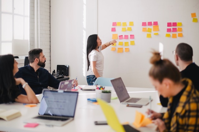 A team meeting with staff sitting around a table in front of a whiteboard covered with post-its. 
