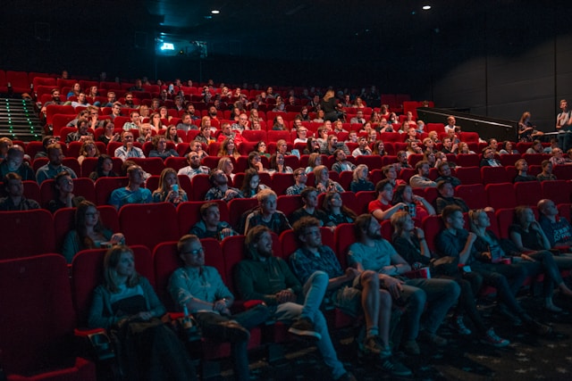 People sit on red chairs in a movie theater. 
