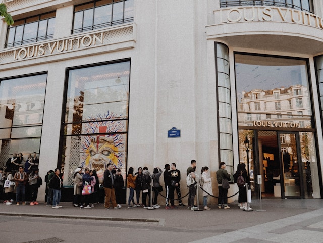 A long line of shoppers waits outside the doors of a luxury store. 
