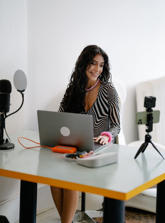 A woman smiles at a mounted tripod while working on her laptop.
