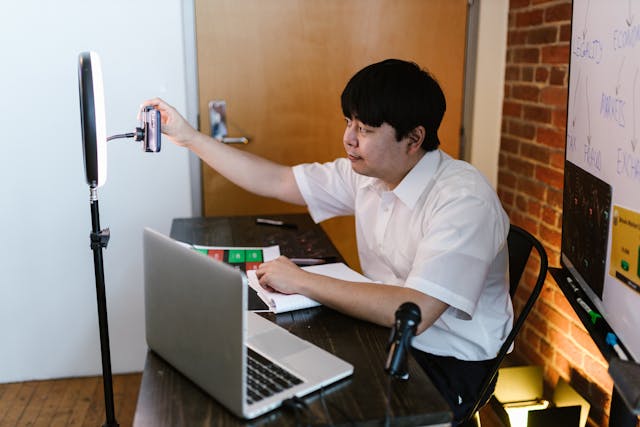 A man at his desk with a laptop adjusts his phone.
