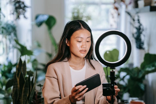 A woman holds her cell phone next to her tripod and ring light.
