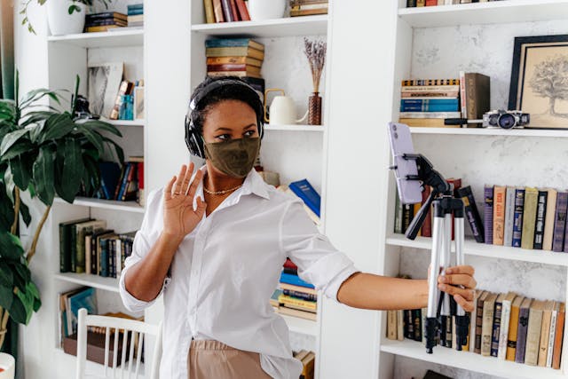 A woman in a mask smiles at her phone on a tripod.
