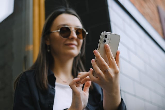 A woman wears sunglasses as she types on her cell phone.
