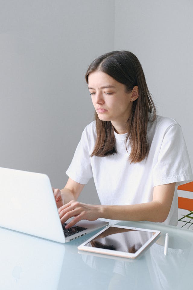 A woman in a white t-shirt types on her laptop beside her tablet.
