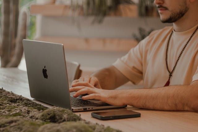 A man sits at a desk to use a MacBook.
