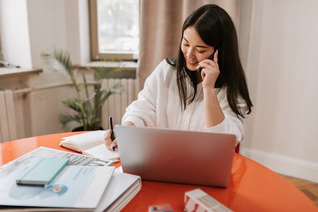 A woman writes as she talks on her phone behind a laptop.
