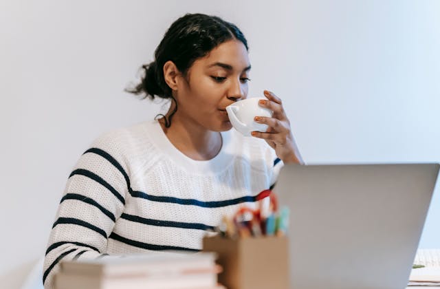 A woman sips her coffee while she types on her laptop.
