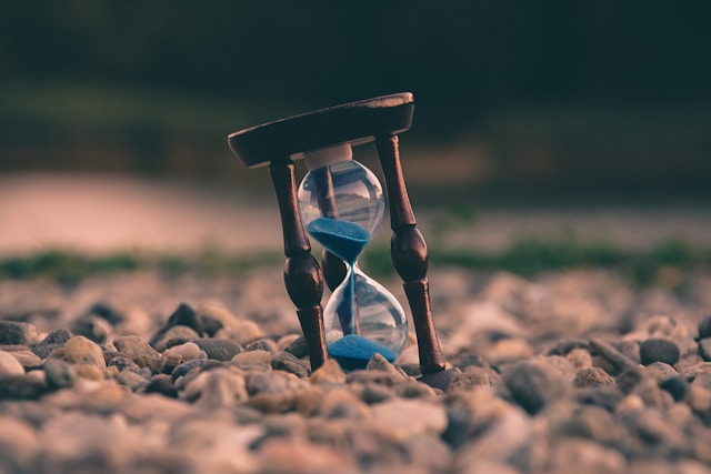 An hourglass timer with blue sand sits on a rocky beach. 
