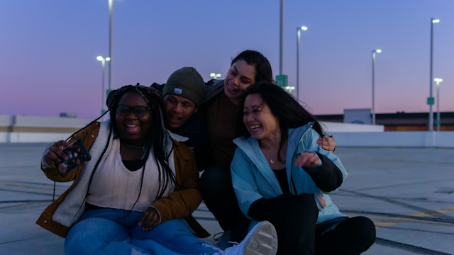 A group of friends on a rooftop takes a group selfie with a phone. 
