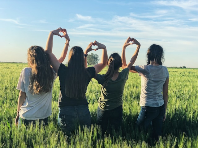 A group of friends stands in a grass field and makes heart shapes with their paired hands. 
