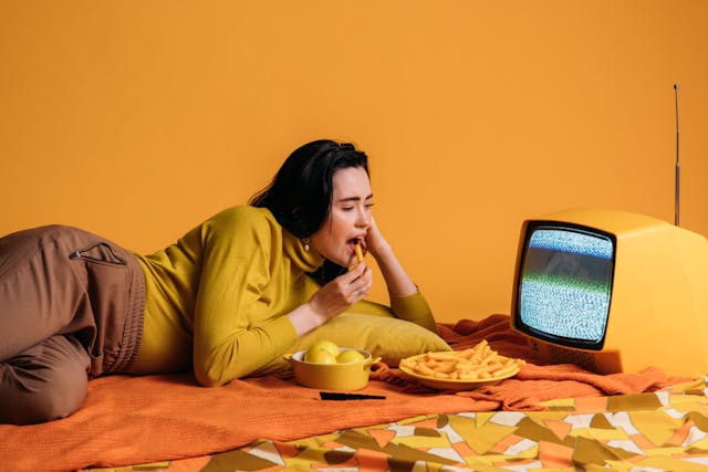 A woman lying down and eating fries while looking at the TV.