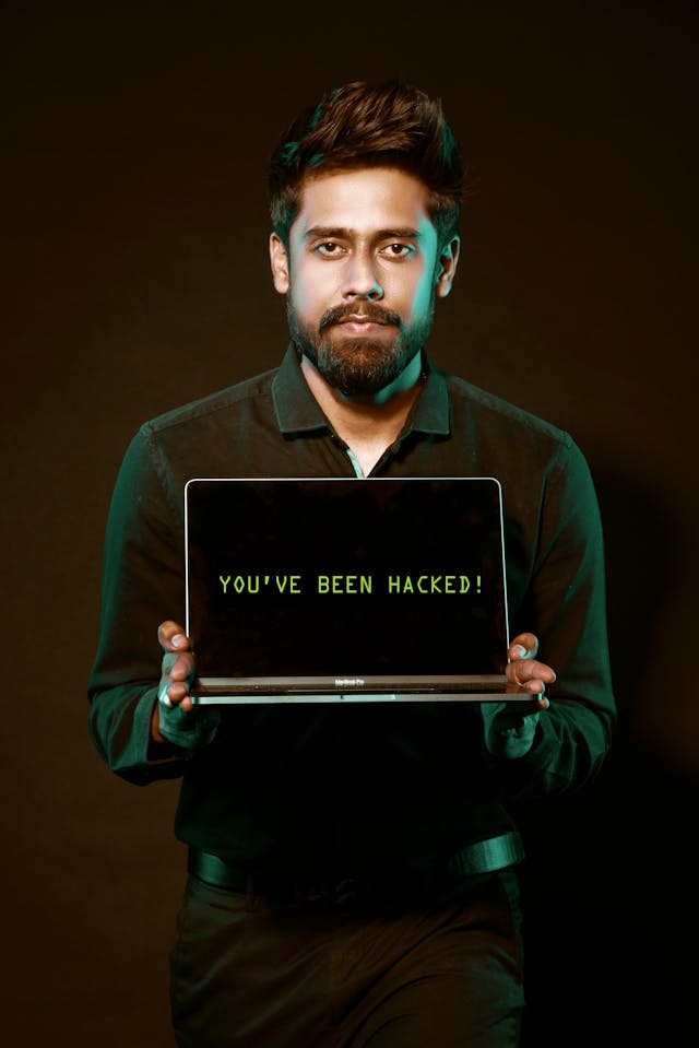 A man with a straight face holding a laptop with “You’ve been hacked!” on the screen.