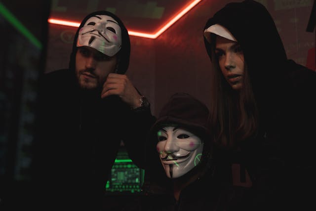 Three people wearing masks and black hoodies hacking a computer system.