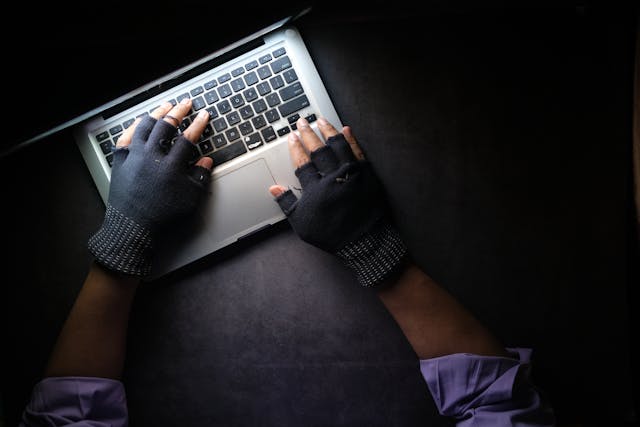 Someone wearing gray fingerless gloves typing on a laptop in a dark room.>