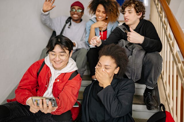 A group of friends sitting on a staircase laughing while watching a video on someone’s phone.