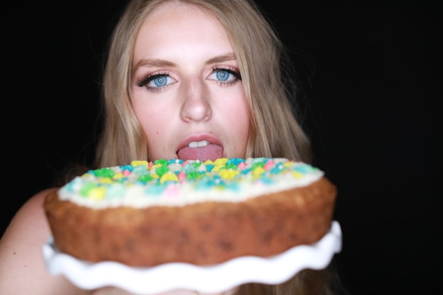 A blonde woman holding up a colorful cake while opening her mouth to take a bite.